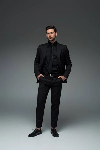 Uomo in elegante abito nero in piedi con le dita nelle tasche sul grigio — Foto stock