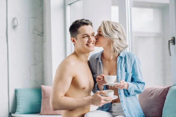 Girlfriend kissing boyfriend and holding cup of coffee at home — Stock Photo