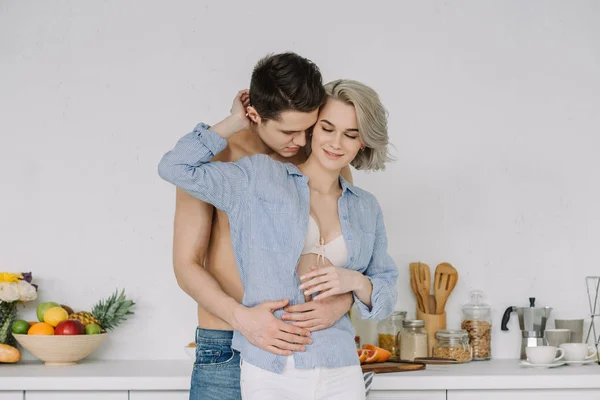 Shirtless boyfriend hugging girlfriend in shirt and bra at kitchen — Stock Photo