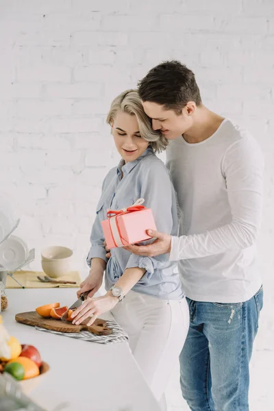 Freundin schneidet Grapefruit und Freund schenkt ihr Geschenkkarton in der Küche, Konzept zum Internationalen Frauentag — Stockfoto