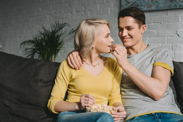 Feliz novio alimentación novia con palomitas de maíz en sofá - foto de stock