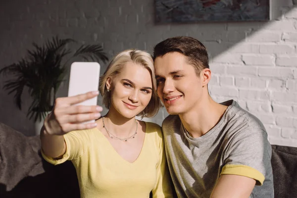 Улыбающаяся пара делает селфи со смартфоном в гостиной — стоковое фото