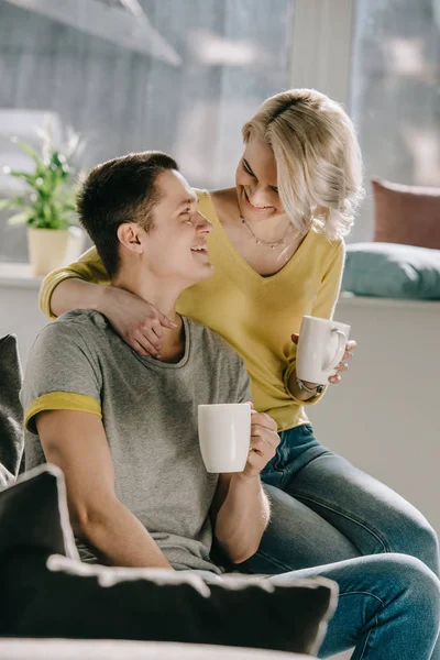 Улыбающаяся пара, держащая чашки кофе и глядя друг на друга дома — стоковое фото