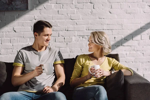 Lächelndes Paar, das Tassen Kaffee hält und sich zu Hause anschaut — Stockfoto