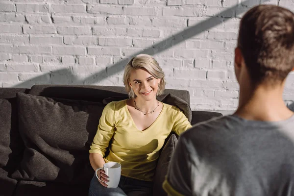 Sonriente novia sosteniendo taza de café y mirando novio en casa - foto de stock