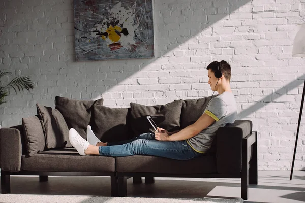 Seitenansicht eines gutaussehenden Mannes, der Musik mit Tablet auf dem Sofa hört — Stockfoto