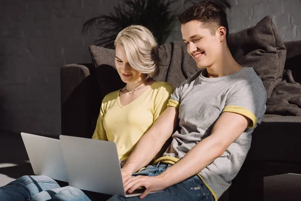 Couple heureux assis sur le sol et en utilisant deux ordinateurs portables à la maison — Photo de stock