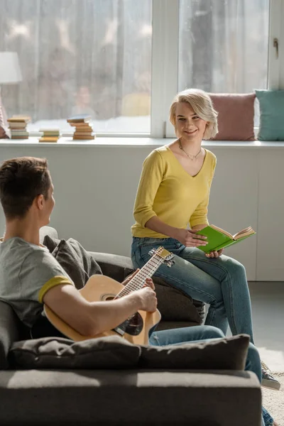 Boyfriend holding acoustic guitar and girlfriend holding book on sofa — Stock Photo