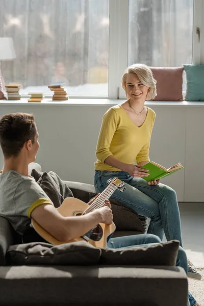 Novio sosteniendo la guitarra acústica y novia sosteniendo libro en el sofá - foto de stock