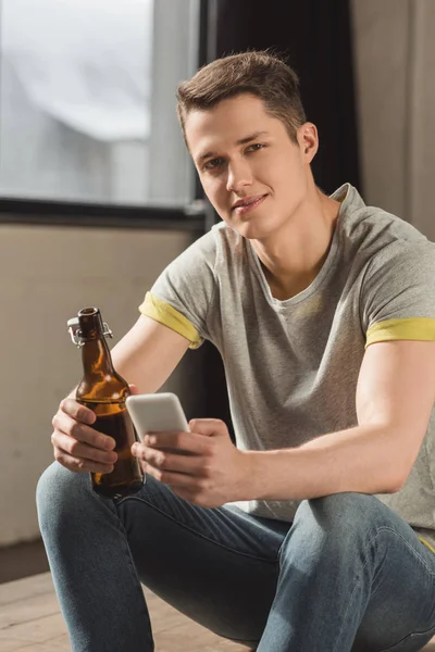 Homem bonito segurando smartphone e garrafa de cerveja e olhando para a câmera em casa — Fotografia de Stock