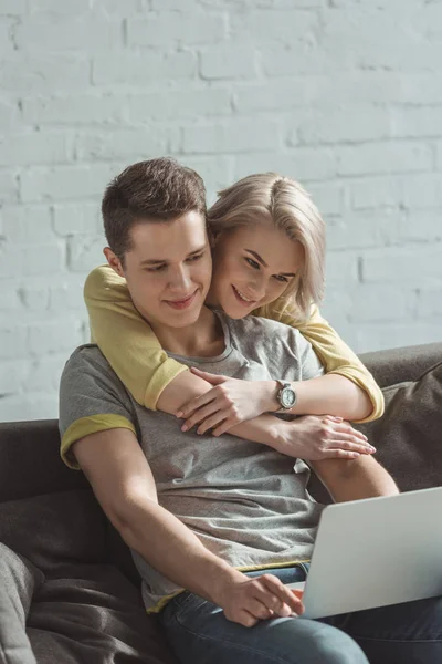 Smiling girlfriend hugging boyfriend and they looking at laptop at home — Stock Photo