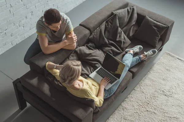 Vista de ángulo alto de la novia usando el ordenador portátil y el novio apoyado en el sofá en casa - foto de stock