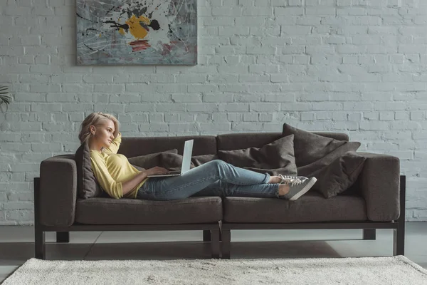 Vue latérale de belle fille couchée avec un ordinateur portable sur le canapé — Photo de stock