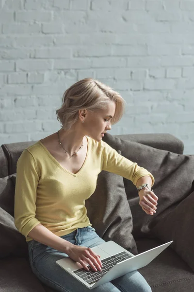 Hermosa chica sosteniendo portátil y mirando el reloj en el sofá - foto de stock