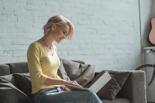 Side view of smiling girl sitting with laptop on sofa — Stock Photo