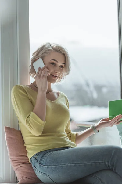 Hermosa chica sosteniendo libro y hablando por teléfono inteligente - foto de stock