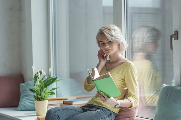 Beautiful girl sitting on windowsill and reading book — Stock Photo