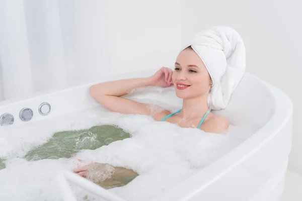 Mulher sorridente com toalha na cabeça relaxante no banho com espuma no salão de spa — Fotografia de Stock