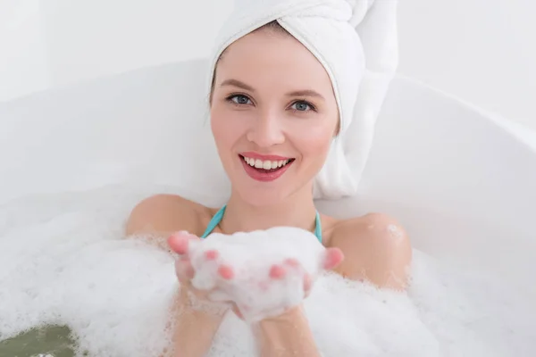 Усміхнена жінка з рушником на голові розслабляється у ванні з піною в спа-салоні — стокове фото