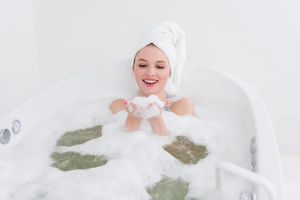 Donna sorridente con asciugamano sulla testa rilassante in bagno con schiuma nel salone spa — Foto stock