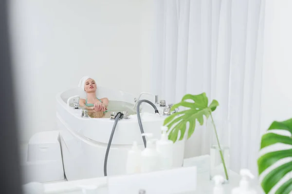 Jeune femme en maillot de bain avec serviette sur la tête relaxant dans la baignoire dans le salon de spa — Photo de stock