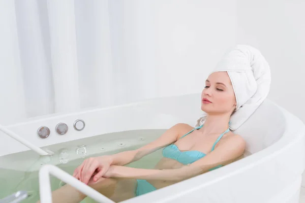 Jeune femme en maillot de bain avec serviette sur la tête relaxant dans la baignoire dans le salon de spa — Photo de stock