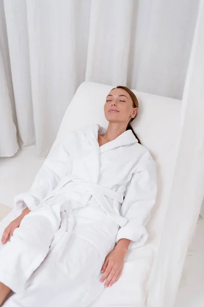 Mujer atractiva joven en albornoz blanco con los ojos cerrados descansando en el salón de spa — Stock Photo
