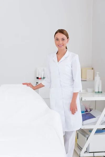 Retrato de cosmetologista sorridente em casaco branco em pé na mesa de massagem vazia no salão — Fotografia de Stock