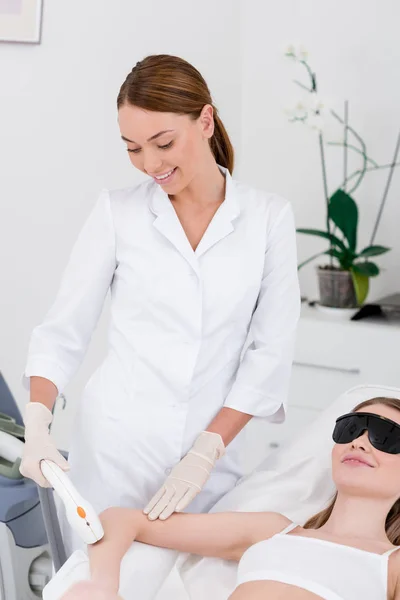 Woman receiving laser hair removal procedure on arm made by cosmetologist in salon — Stock Photo