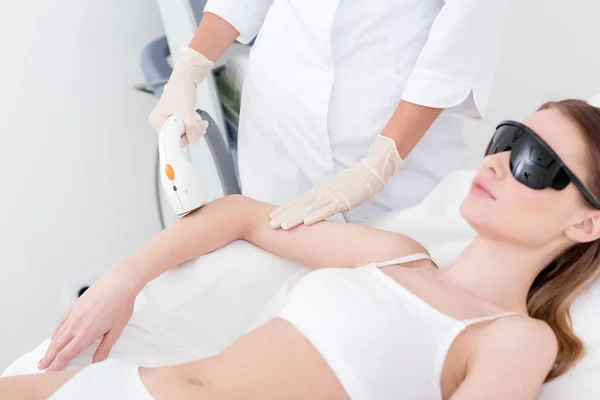 Vista parcial de la mujer que recibe el procedimiento de depilación láser en el brazo realizado por el cosmetólogo en el salón - foto de stock