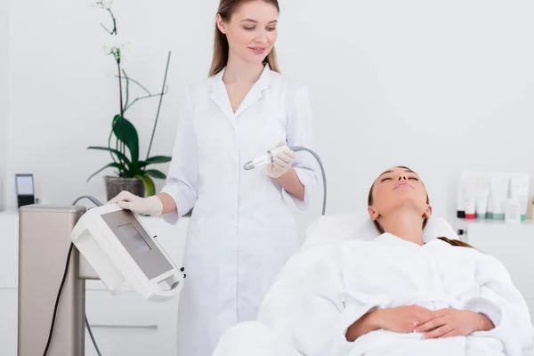 Jeune femme couchée sur une table de massage dans un salon de cosmétologie avec un cosmétologue proche — Photo de stock