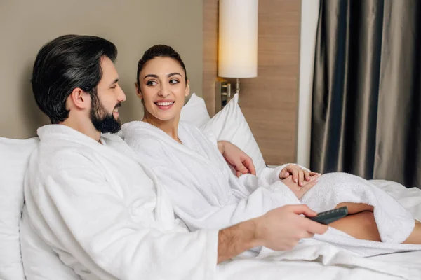 Happy couple in bathrobes with tv remote control relaxing in bed at hotel suite — Stock Photo