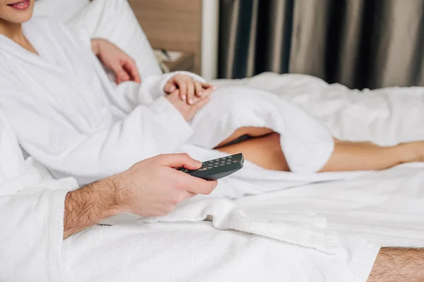 Tiro recortado de pareja en albornoces con control remoto de televisión relajante en la cama en la suite del hotel - foto de stock
