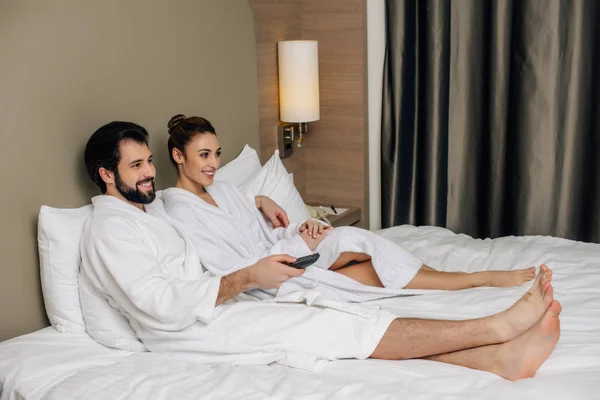 Beautiful couple in bathrobes with remote control relaxing in bed at hotel suite and watching tv — Stock Photo
