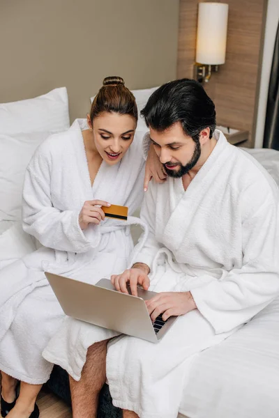 Belo casal em roupões de banho fazendo e-shopping com laptop e cartão de crédito na suíte do hotel — Fotografia de Stock