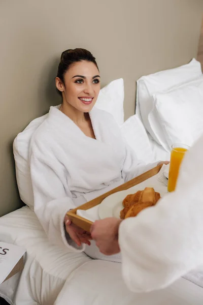 Bela mulher em roupão levando comida na bandeja do homem enquanto relaxa na cama — Fotografia de Stock