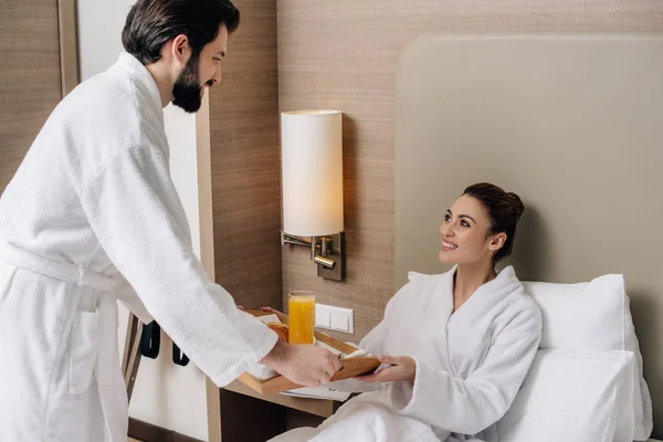Man giving tray with breakfast to girlfriend while she sitting in bed at hotel suite — Stock Photo