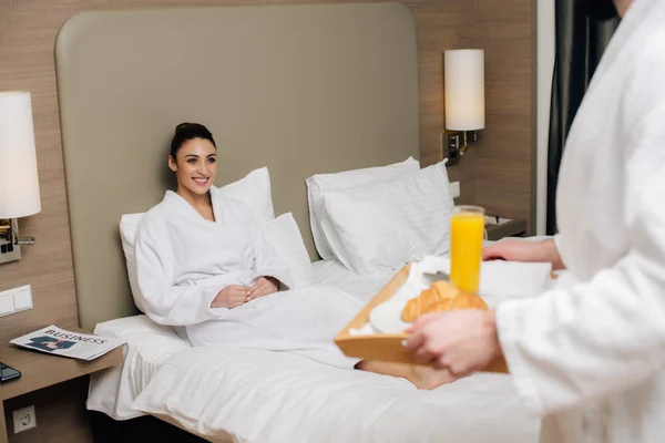 Abgeschnittene Aufnahme von Mann, der Tablett mit leckerem Frühstück für Freundin trägt, während sie in der Hotelsuite im Bett sitzt — Stockfoto