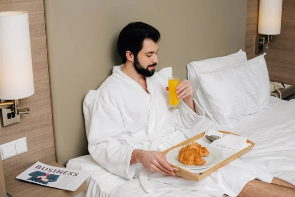 Homem bonito em roupão de banho com croissants e suco na bandeja sentado na cama na suíte do hotel — Fotografia de Stock