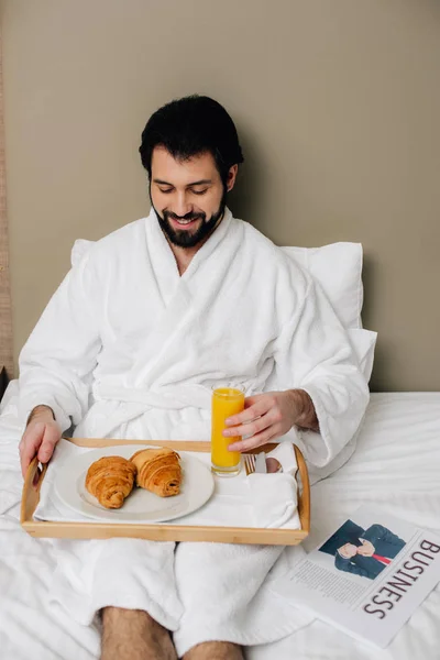 Homem sorridente em roupão de banho com comida na bandeja sentado na cama na suíte do hotel — Fotografia de Stock