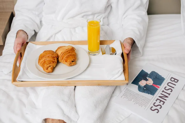 Обрезанный снимок мужчины в халате с едой на подносе, сидящего на кровати в номере отеля — стоковое фото