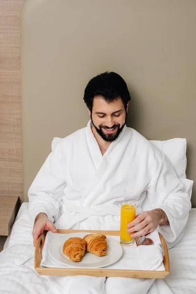 Homem feliz em roupão de banho com comida na bandeja sentado na cama na suíte do hotel — Fotografia de Stock