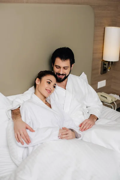 Couple heureux en peignoirs embrassant dans le lit de la suite de l'hôtel — Photo de stock