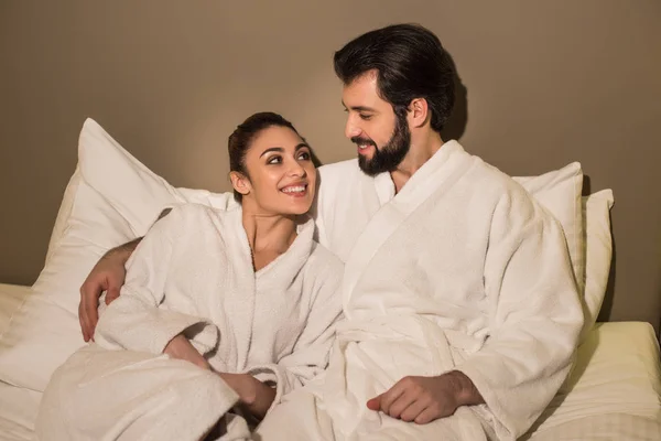 Beautiful couple in bathrobes embracing in bed of hotel suite and looking at each other — Stock Photo