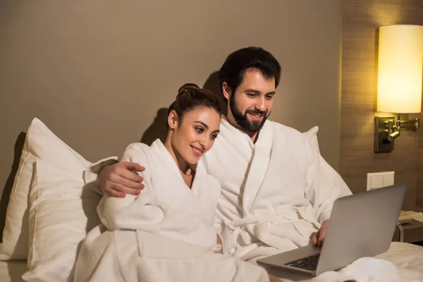 Hermosa pareja en albornoces usando el ordenador portátil juntos en la cama de la suite del hotel - foto de stock