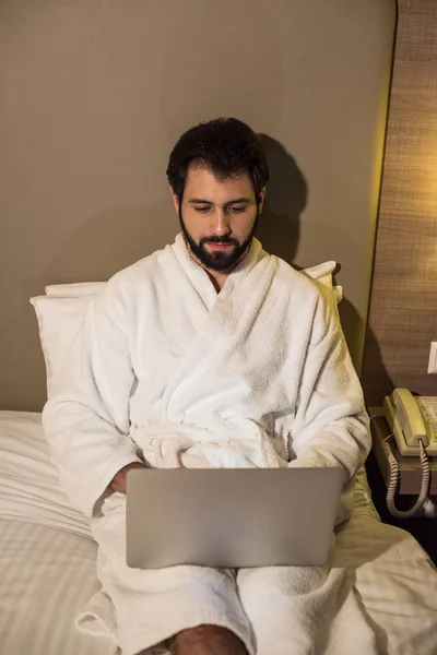 Homem bonito em roupão de banho usando laptop na cama na suíte do hotel à noite — Fotografia de Stock