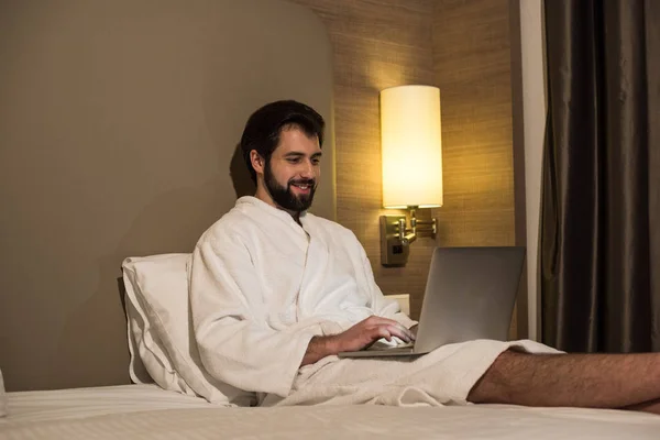 Homme souriant en peignoir en utilisant un ordinateur portable au lit dans la suite de l'hôtel le soir — Photo de stock