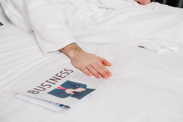 Recortado tiro de hombre en albornoz acostado en la cama con periódico de negocios - foto de stock