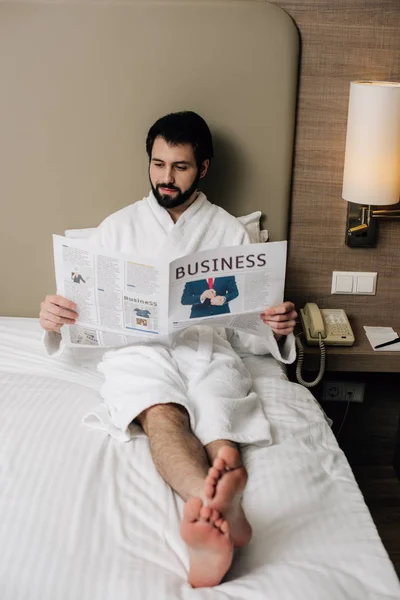 Hombre de negocios descalzo en el periódico de lectura de albornoz mientras se relaja en la cama en la suite del hotel - foto de stock