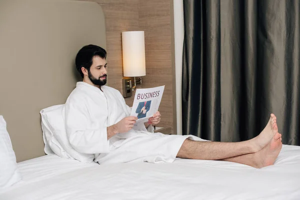 Homme d'affaires souriant en peignoir lisant le journal tout en se relaxant au lit dans la suite de l'hôtel — Photo de stock